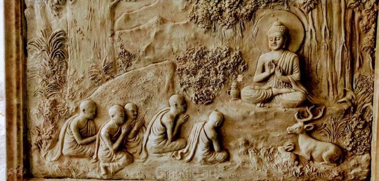 A deep fiber cast relief mural of the Buddha teaching beneath trees, with disciples in the foreground and deer at the edge of the composition